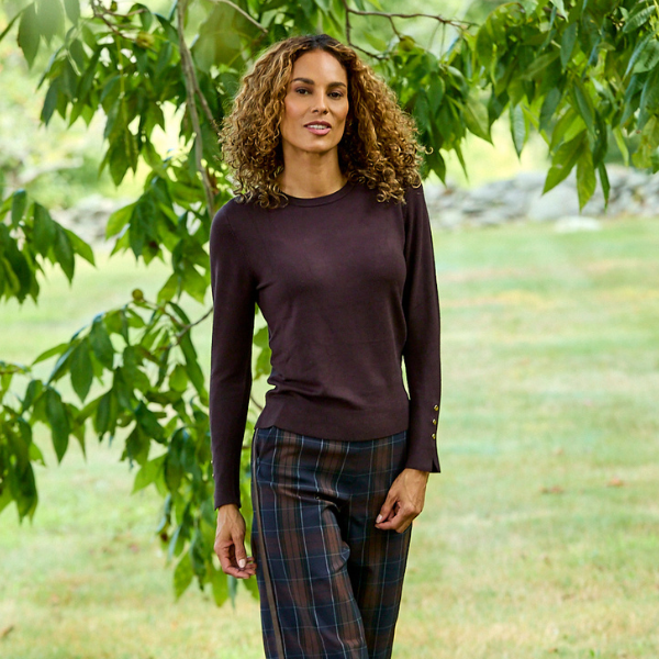 A woman with curly brown hair stands outdoors in a grassy park area under leafy trees, wearing a dark plum long-sleeve knit top and dark plaid pants.