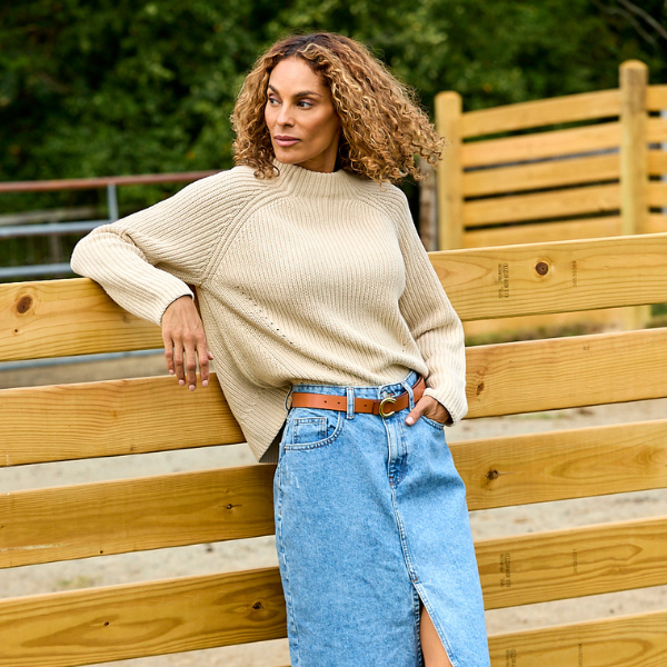A woman with curly brown hair leans against a wooden fence outdoors, wearing a cream ribbed knit mock-neck sweater and a light blue denim skirt with a brown belt.