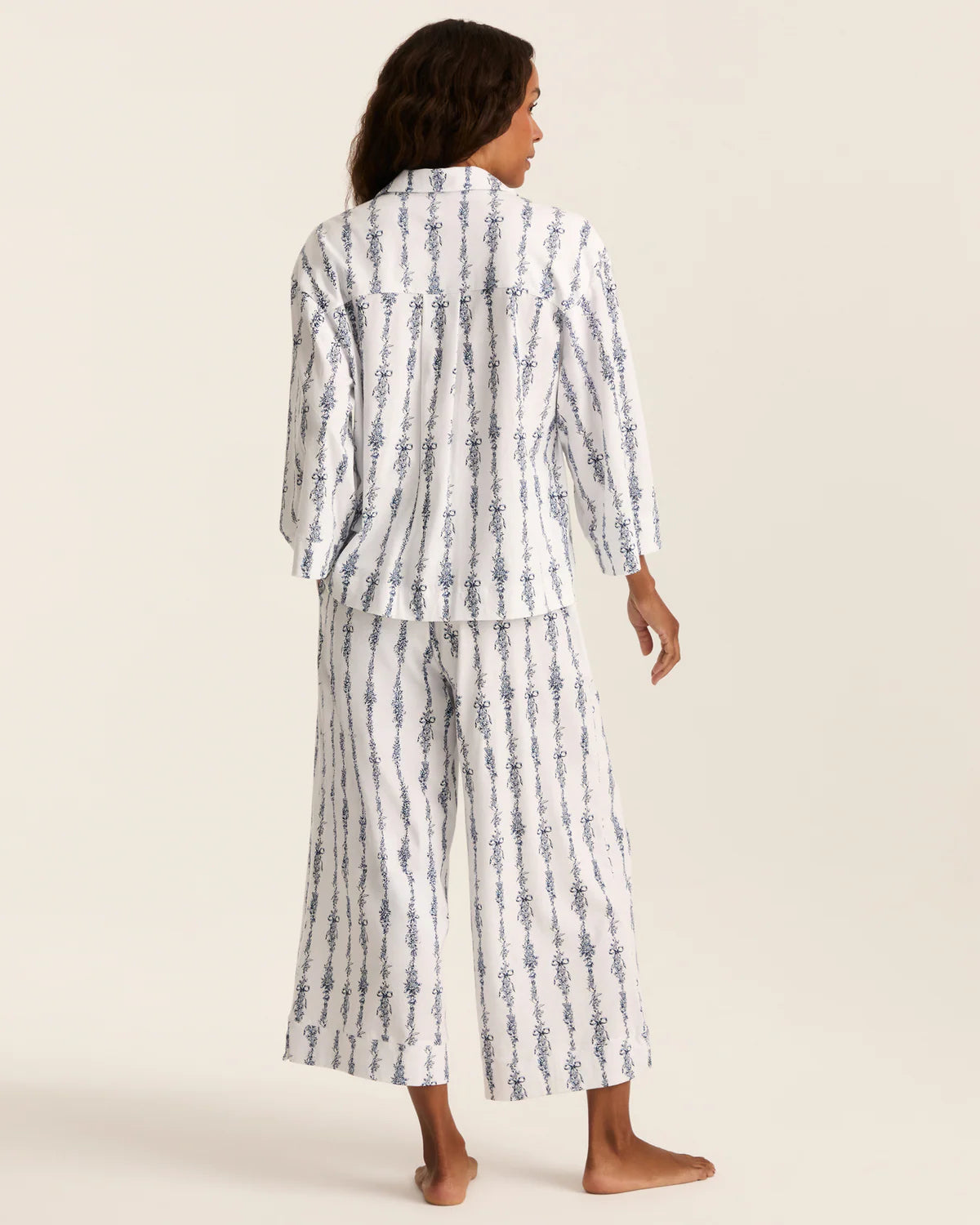 A woman modeling a matching two-piece pajama set with a white base and vertical blue patterned stripes, shown from the back against a solid light beige background.