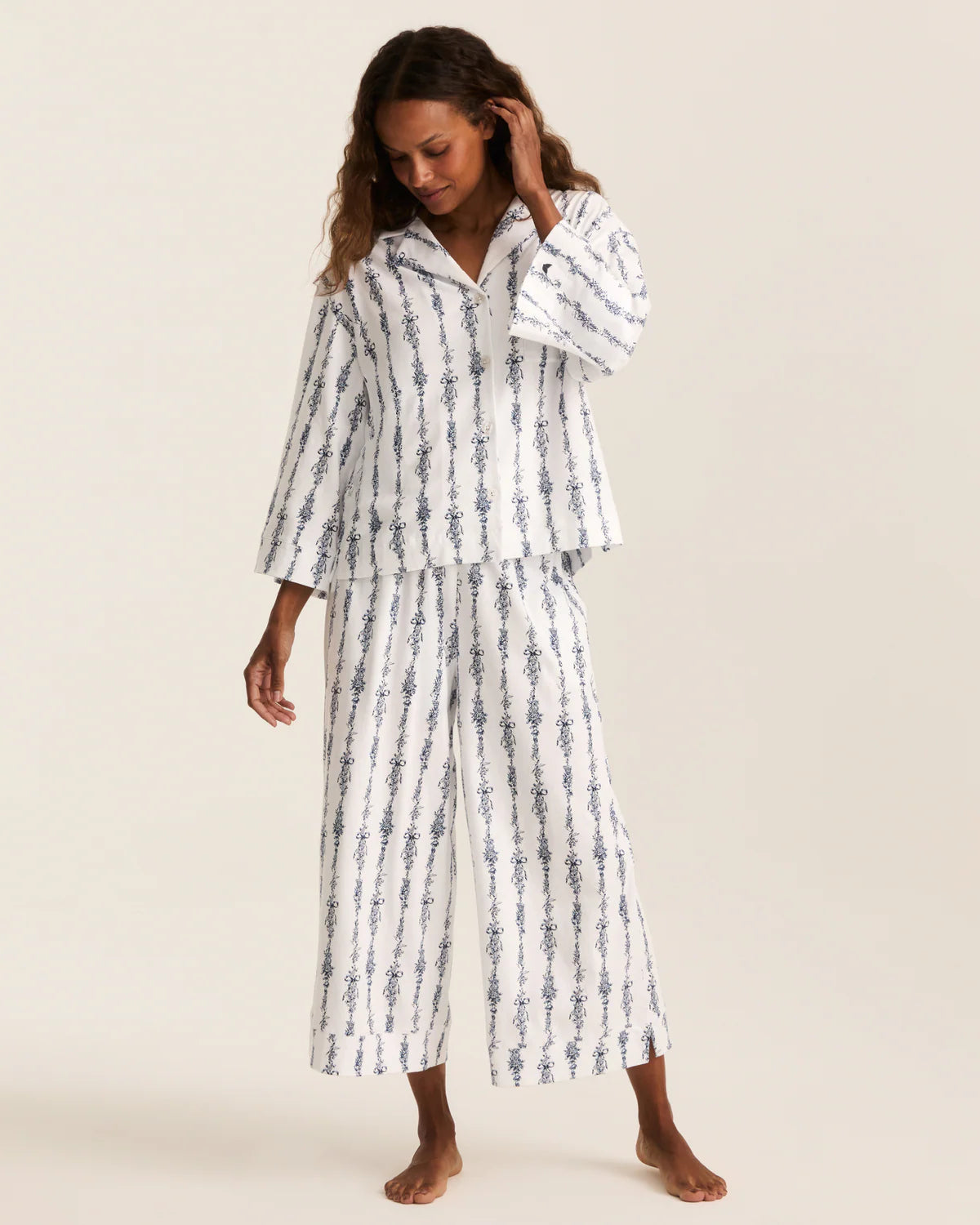 A woman modeling a two-piece pajama set with a white base and vertical blue patterned stripes, featuring a long-sleeve button-up top and matching wide-leg pants, set against a solid light beige background.