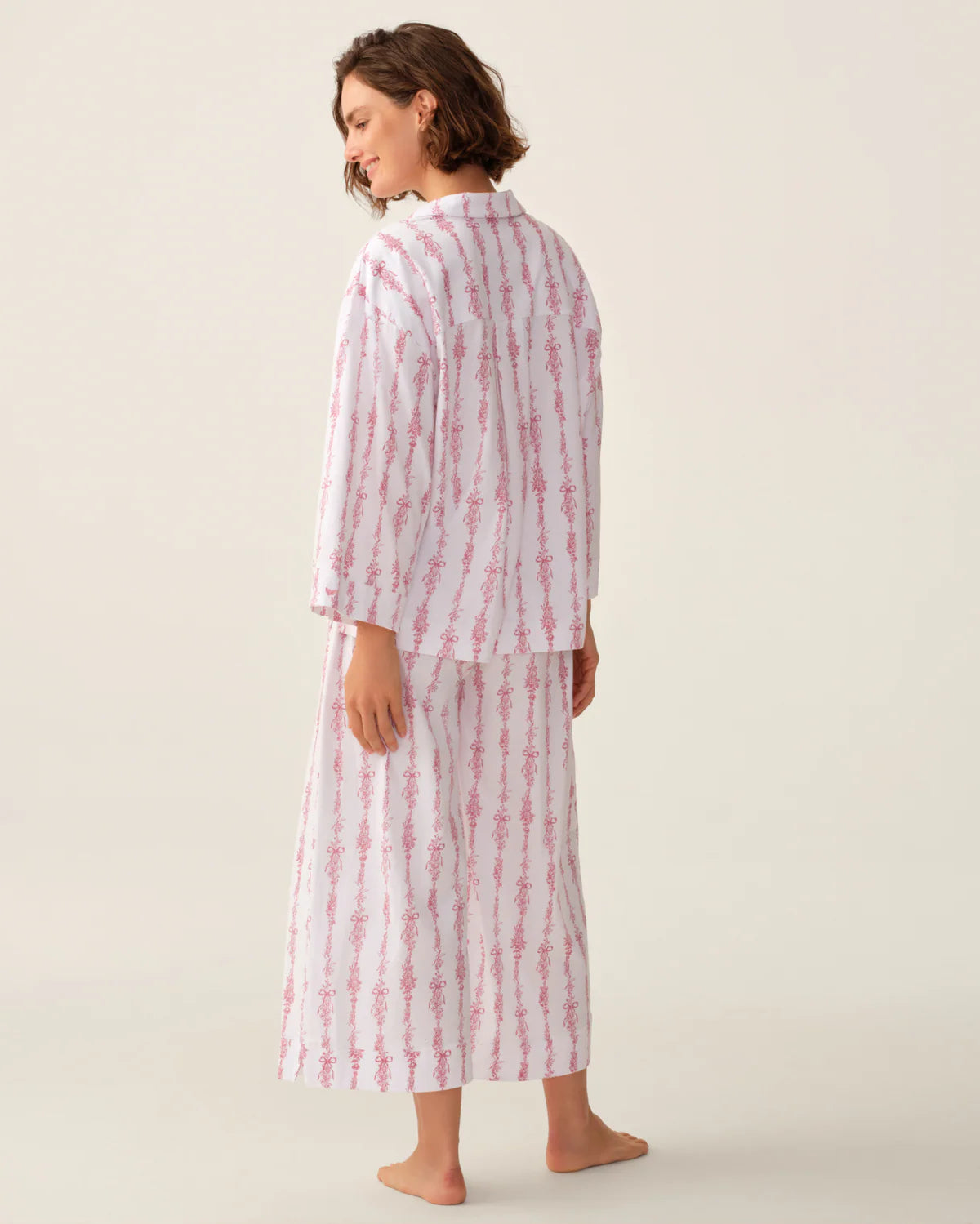 A woman wearing a matching two-piece pajama set with vertical pink and white stripes, shown from a rear three-quarter angle against a plain light beige studio background.