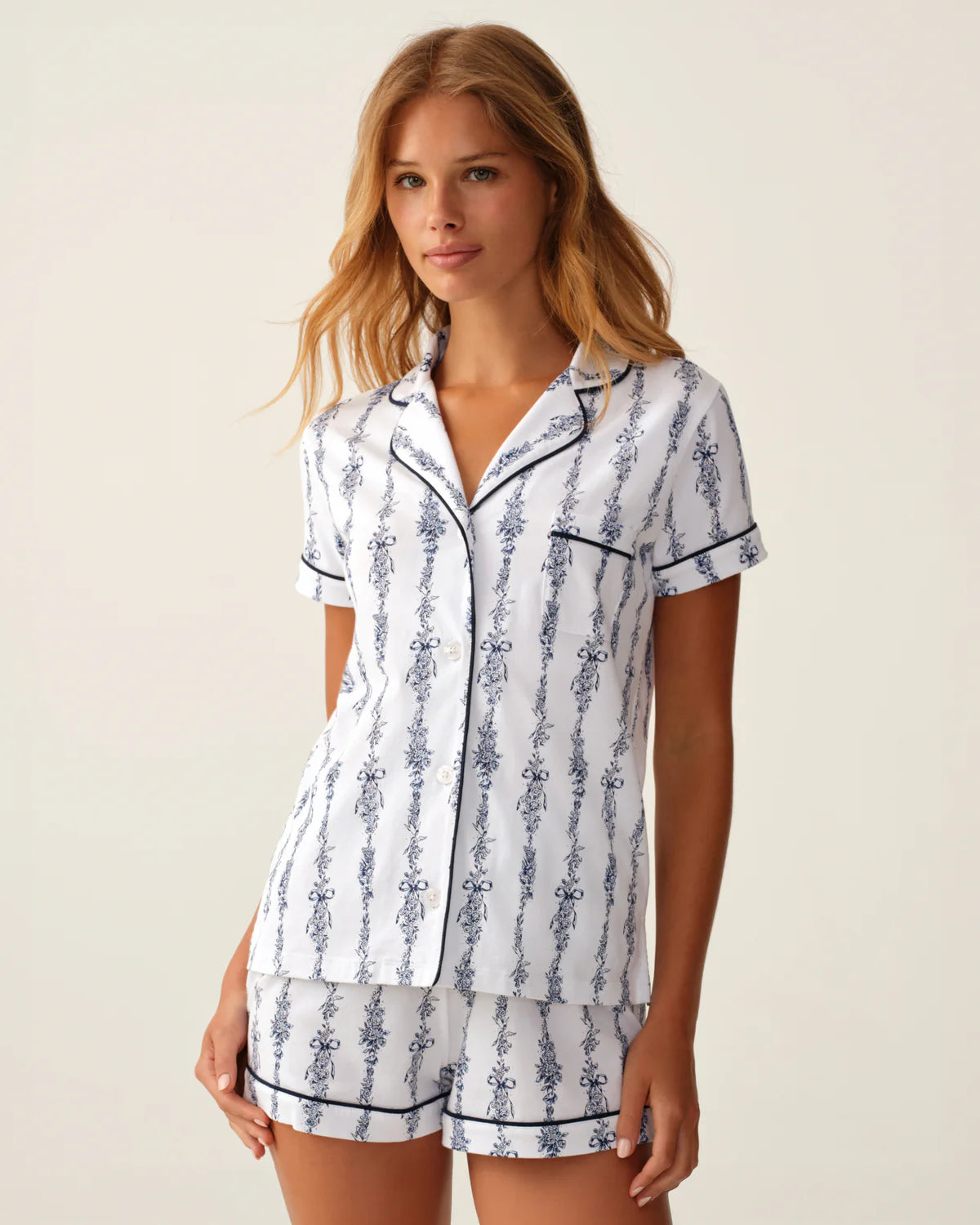 A woman modeling a matching short-sleeve pajama shirt and shorts set with a white base, blue vertical ornamental pattern, and dark piping trim, against a plain light beige studio background.