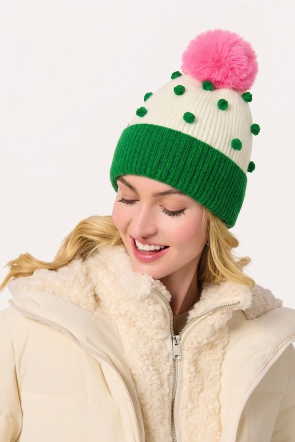 A woman wearing a cactus-themed knit beanie with a pink pom-pom and green cactus details, paired with a cream sherpa-lined winter jacket against a plain white background.
