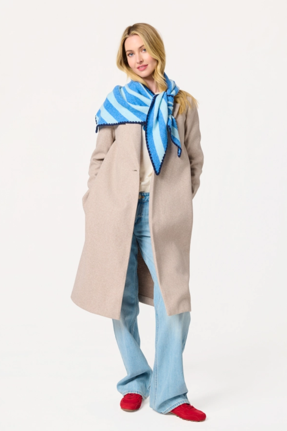 A woman modeling a long beige coat, light blue jeans, a blue and white striped knit scarf, and red slip-on shoes against a plain white studio background.