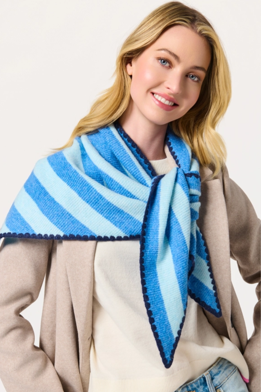A smiling blonde woman wearing a blue and light blue striped knit scarf with dark blue pom-pom trim, styled over a beige cardigan and cream top.