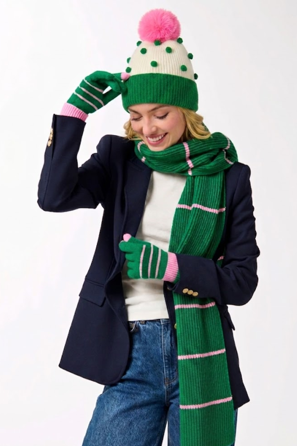 A woman wearing a green and cream knit beanie with a pink pom-pom, matching green striped knit gloves, and a green striped knit scarf, styled with a navy blazer, white top, and blue jeans against a plain white background.