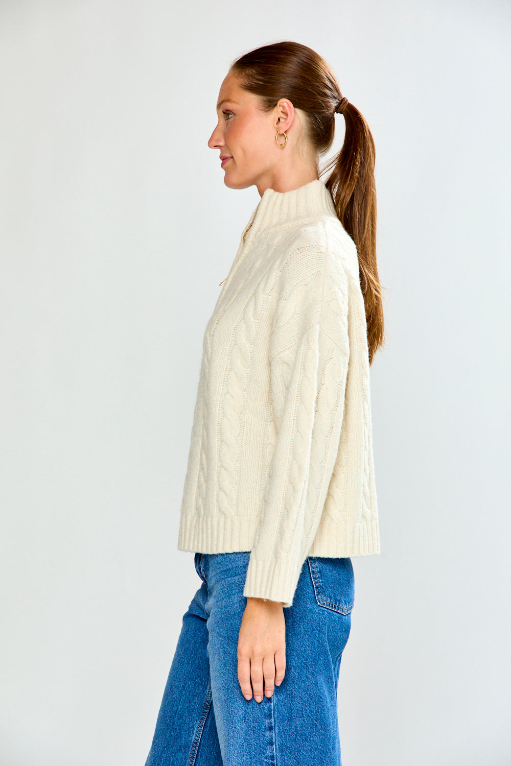 Woman wearing a cream cable knit sweater and blue jeans on a white background