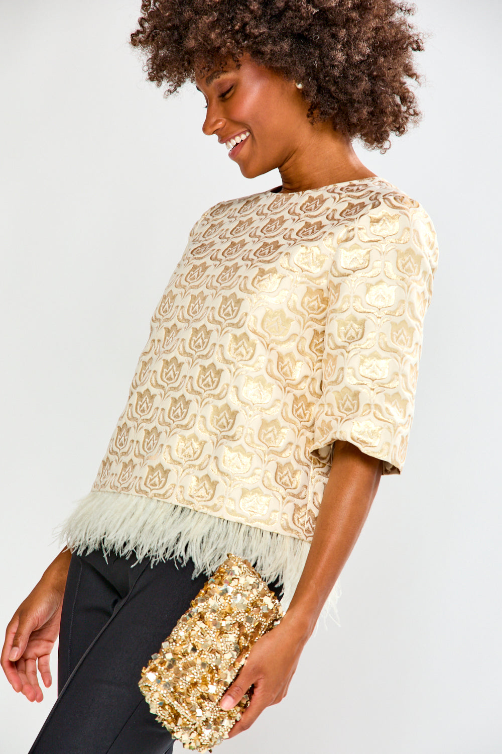 Woman wearing a beige lace top with feather details and holding a gold clutch against a white background