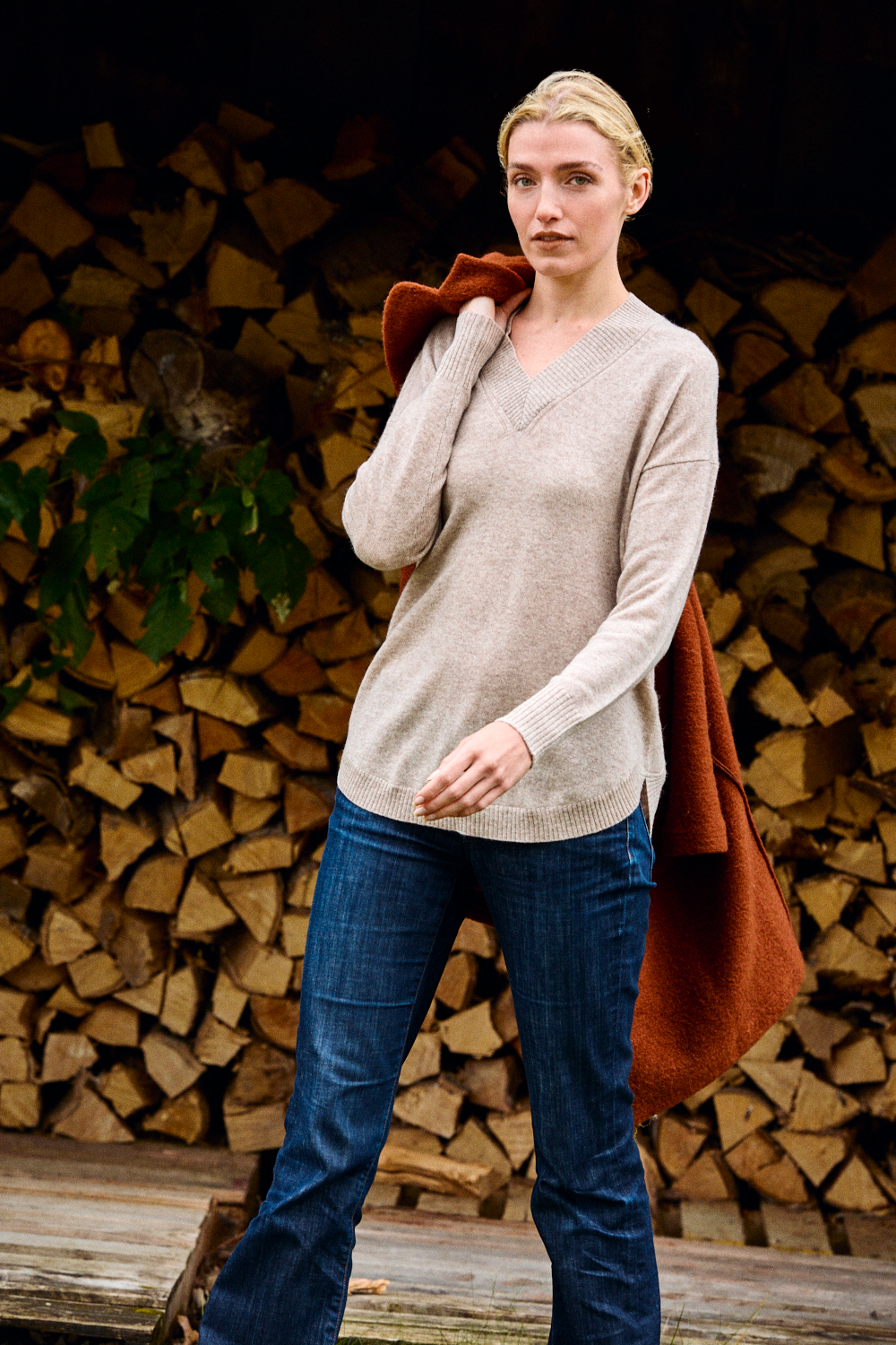 Woman wearing a beige sweater and blue jeans standing in front of stacked firewood.