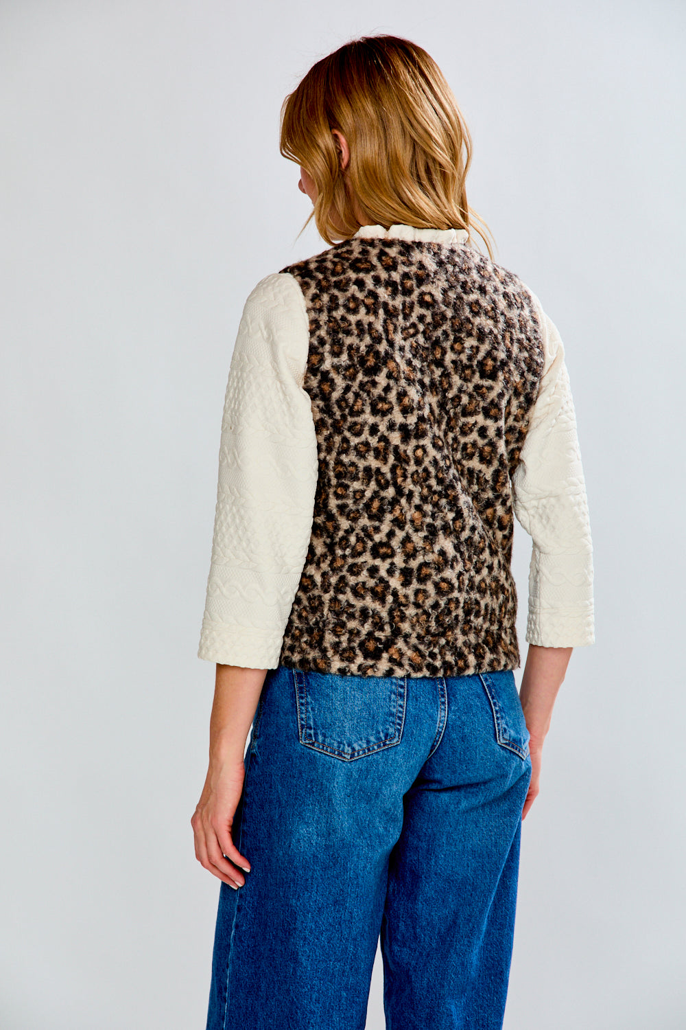 Person wearing a leopard print vest over a white shirt and blue jeans on a plain background