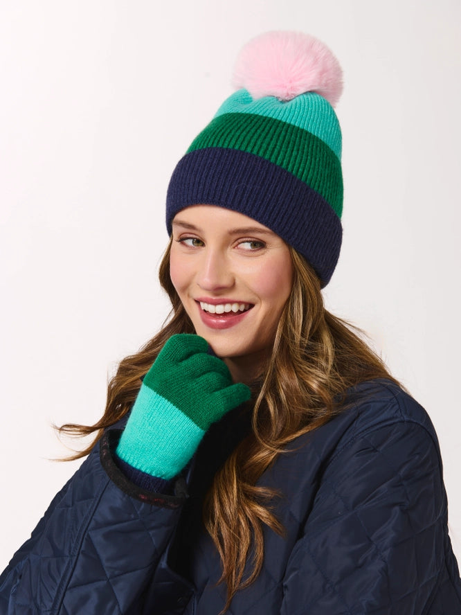 A smiling woman wearing a striped knit beanie with a pink pom-pom, teal and green stripes, and a navy ribbed bottom, paired with matching teal and green knit gloves and a navy quilted jacket, against a plain white background.