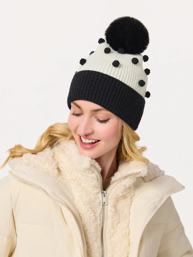 A woman wearing a cream and black knit pom-pom beanie with black pom-poms, paired with a cream puffer jacket with a sherpa collar, against a plain white background.