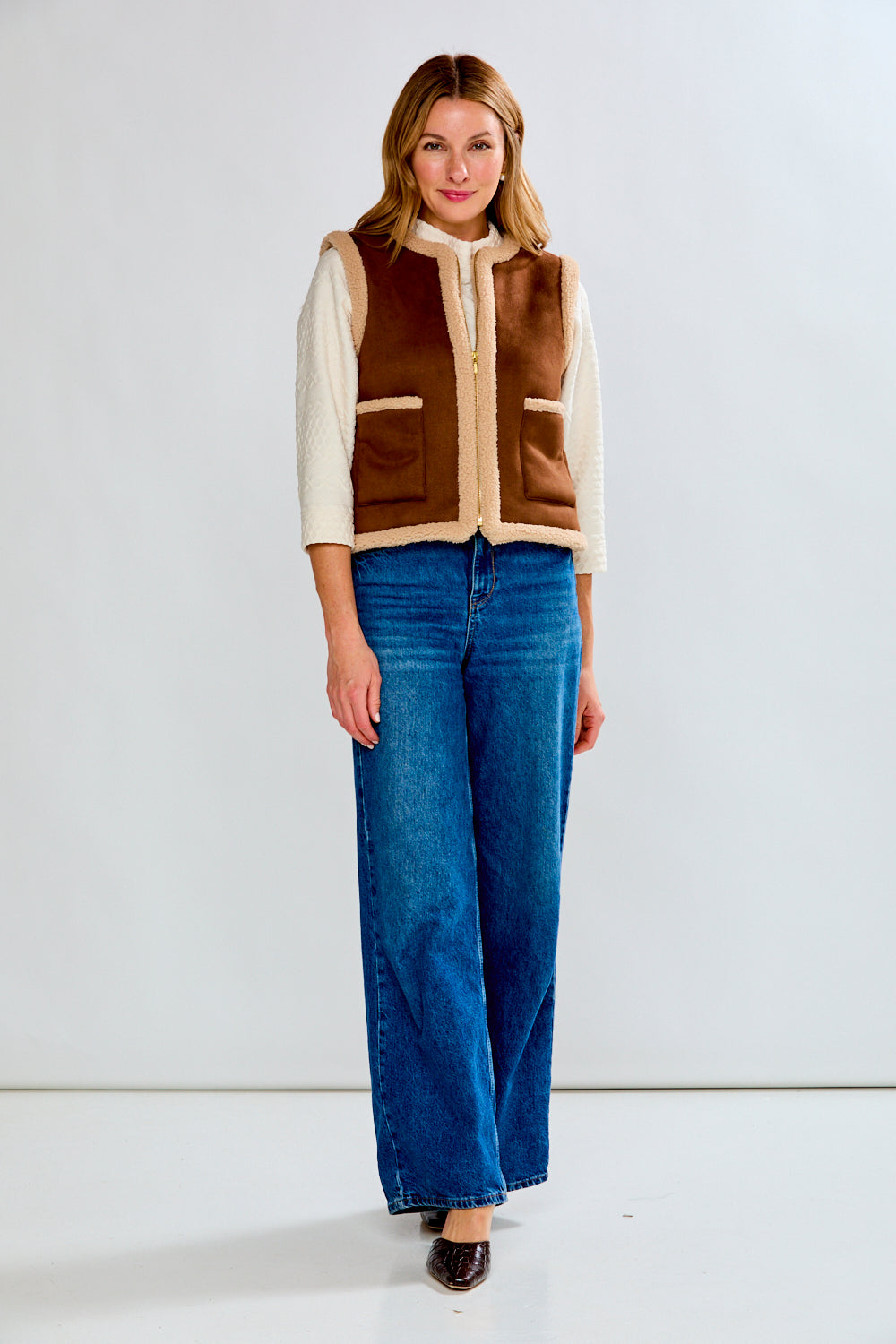 Woman wearing a brown and beige vest over a cream shirt with blue jeans on a white background