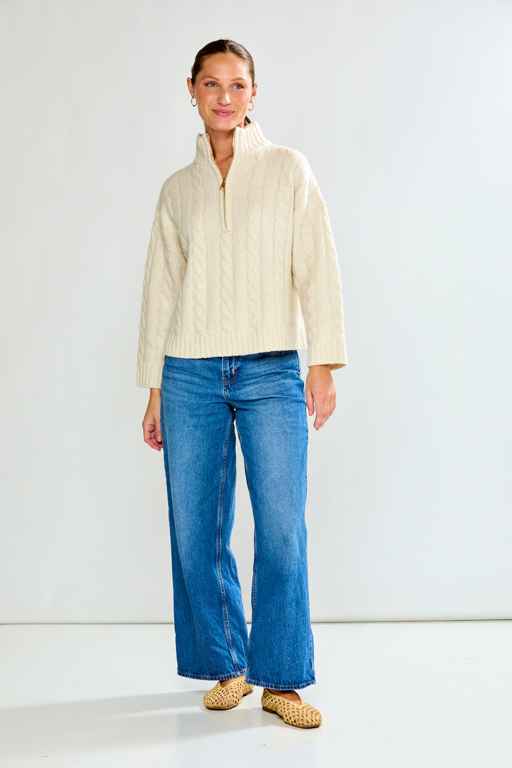 Woman wearing a cream cable knit sweater and blue jeans on a white background