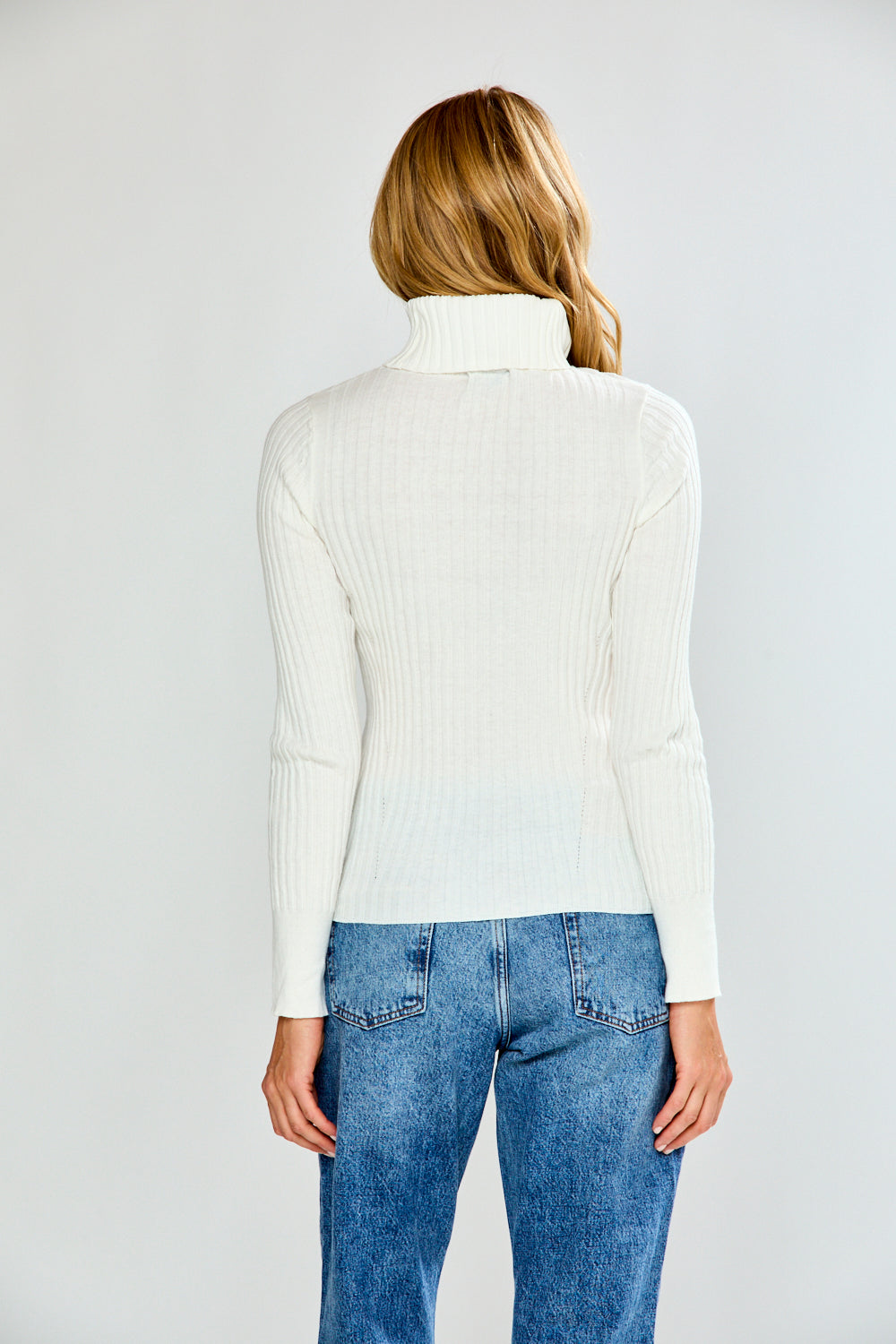 Woman wearing a white turtleneck sweater and blue jeans on a plain background