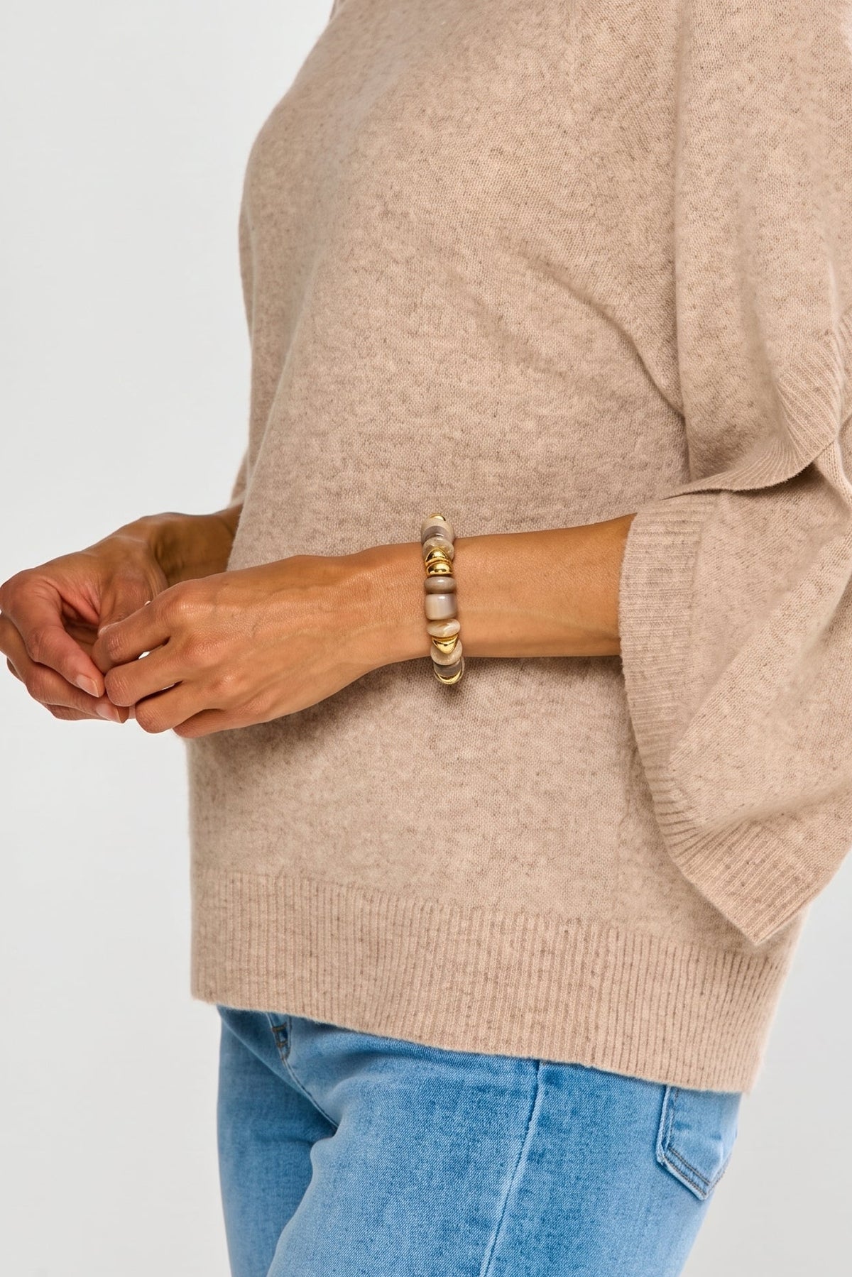 A woman wearing a light beige knit sweater with three-quarter flared sleeves and blue jeans, shown from the side against a plain light gray background.