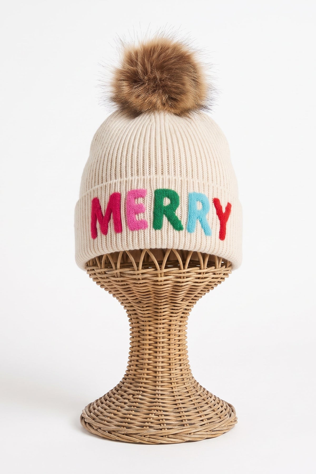 A ribbed knit beanie hat in warm cream with a brown faux-fur pom-pom on top and the word 'MERRY' embroidered across the front in pink, green, and red. The hat is displayed on a woven wicker stand against a clean white background.