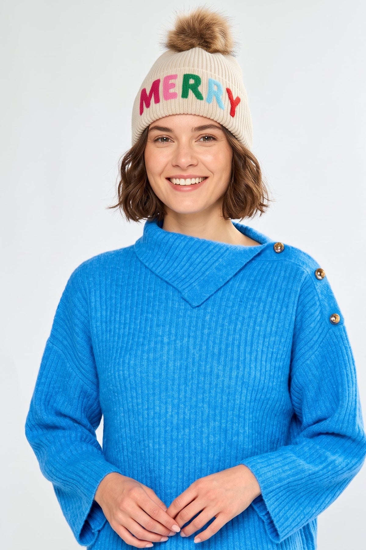A smiling woman wearing a bright blue ribbed knit sweater and a cream knit beanie with a brown pom-pom. The beanie has the word 'MERRY' in colorful letters across the front.