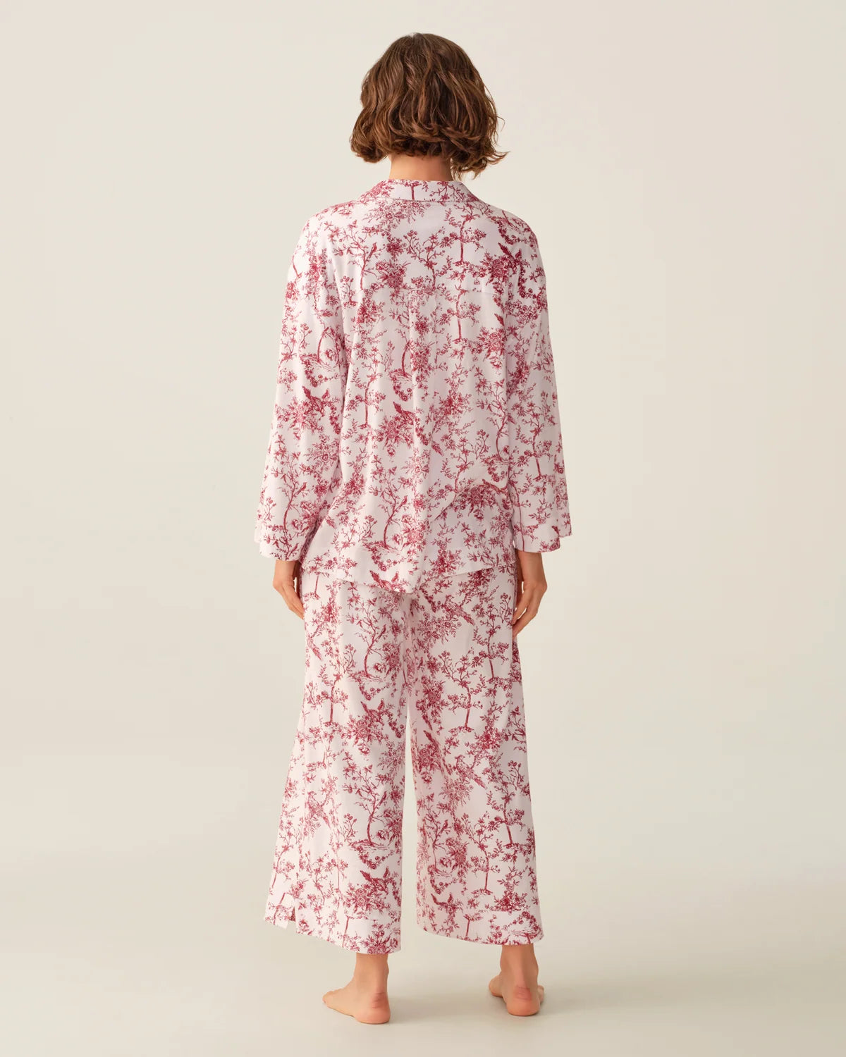 A model viewed from behind wearing a matching two-piece pajama set with a red floral print on a white base, set against a plain light beige studio background.