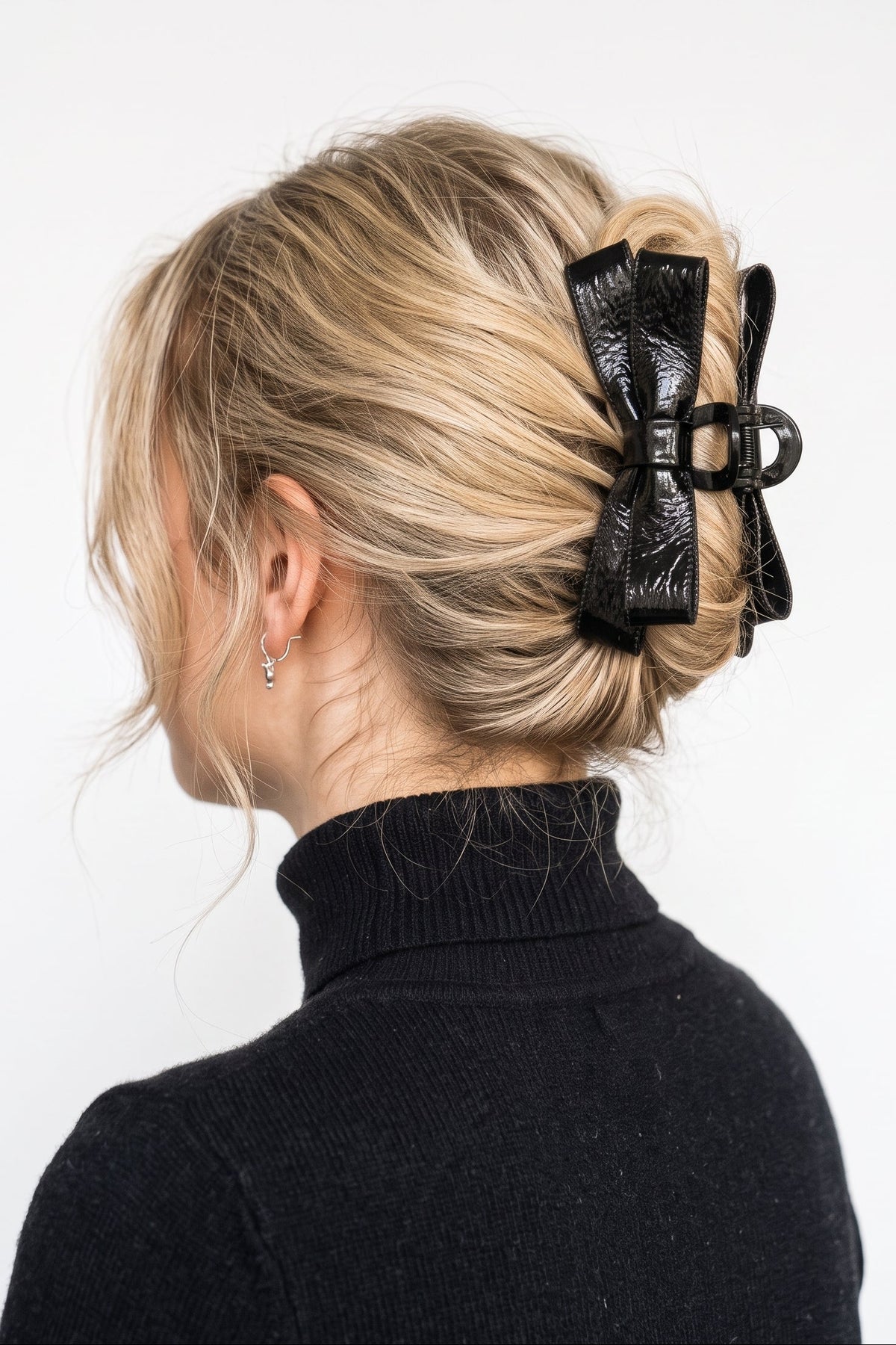 A blonde woman shown from the back wearing a large black textured hair claw clip holding her hair in a low bun, paired with a black turtleneck sweater against a plain white background.
