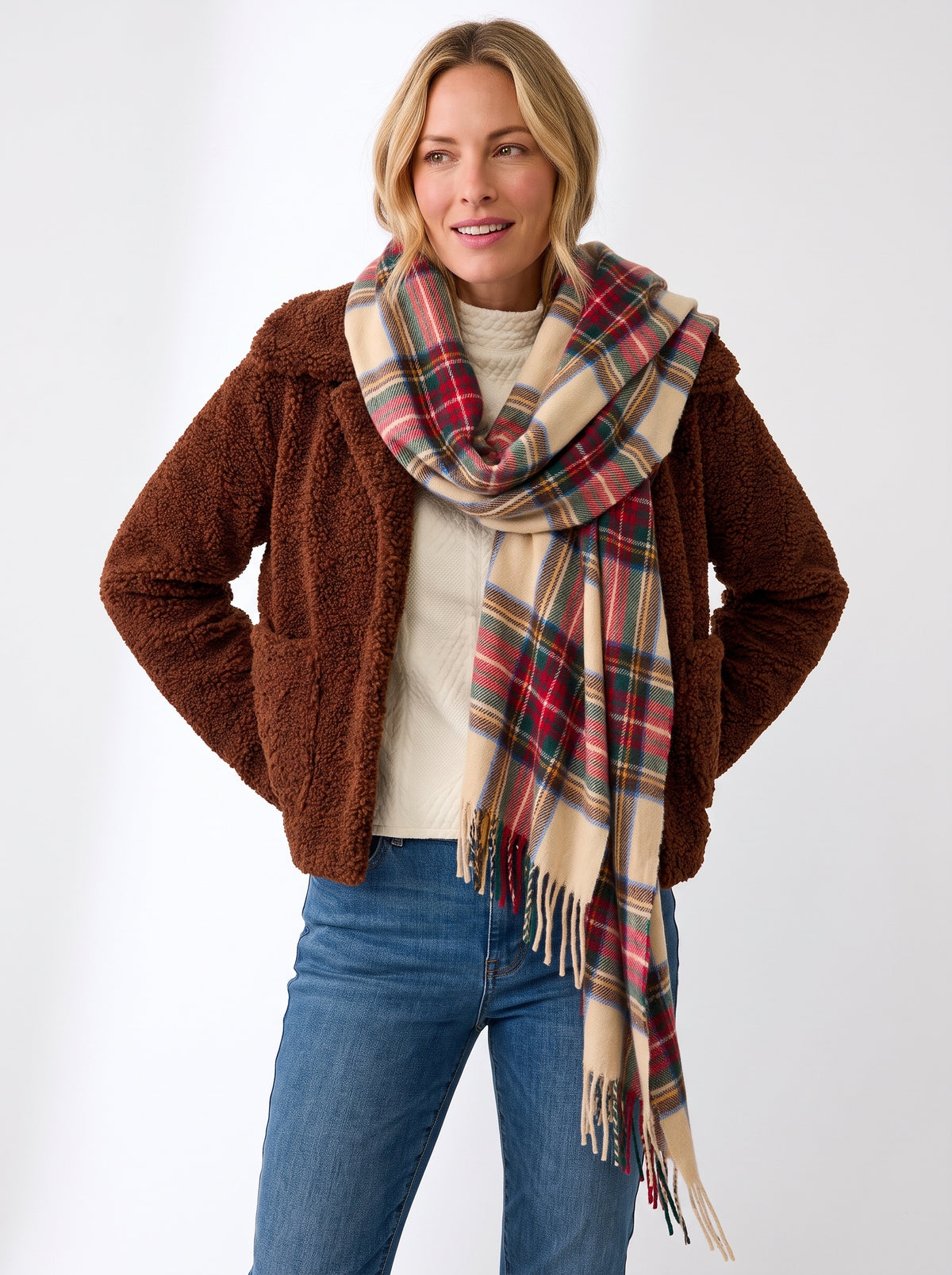 A woman modeling a brown sherpa jacket, cream knit sweater, and a thick plaid scarf in beige, red, green, and blue, paired with blue jeans against a plain white studio background.