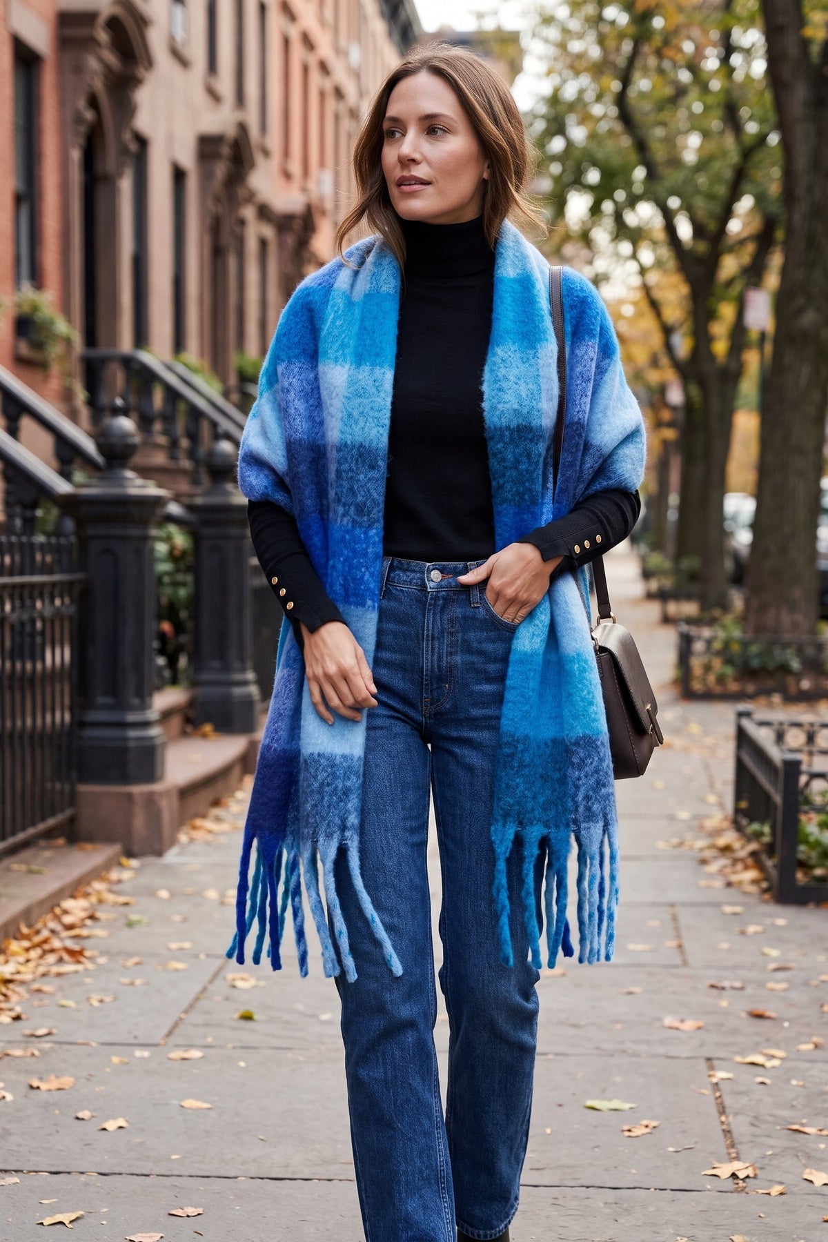 A woman walking on a city sidewalk wearing a blue and turquoise plaid fringed wrap shawl over a black turtleneck and blue jeans.