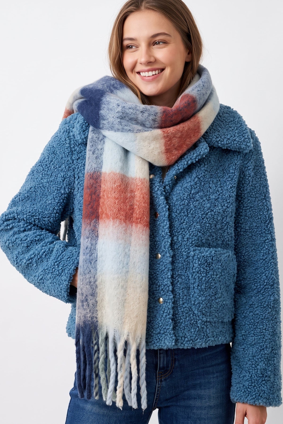 A woman wearing a blue teddy fleece jacket and a thick plaid scarf with blue, white, and rust-orange stripes, posed against a light gray studio background.