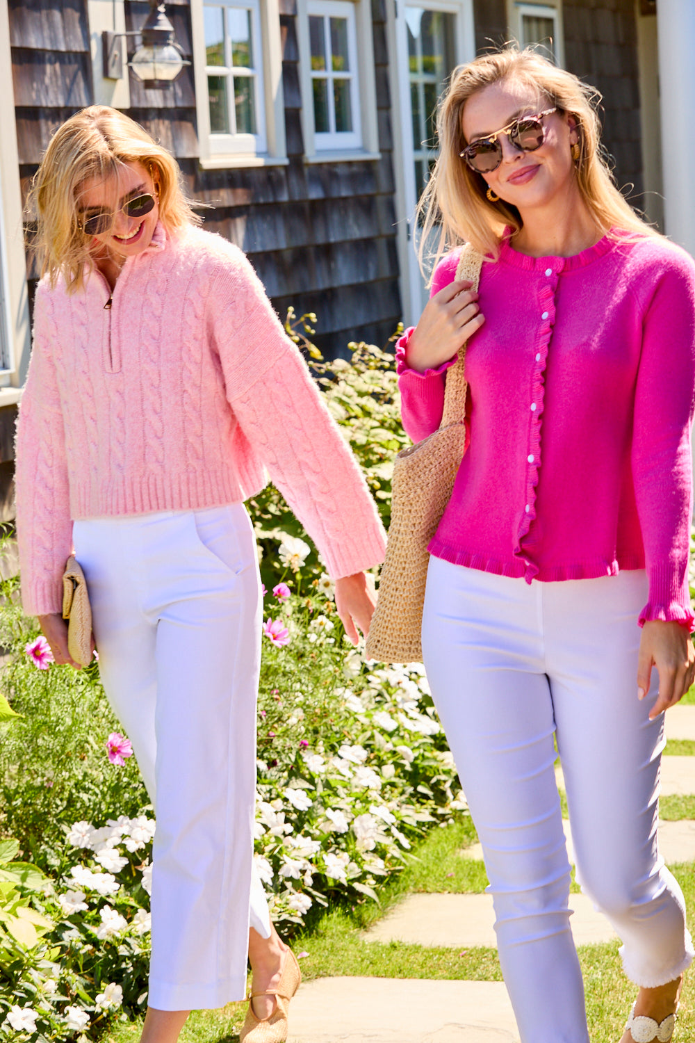 Two women walking outdoors, one in a pink sweater and white pants, the other in a bright pink cardigan and white pants.