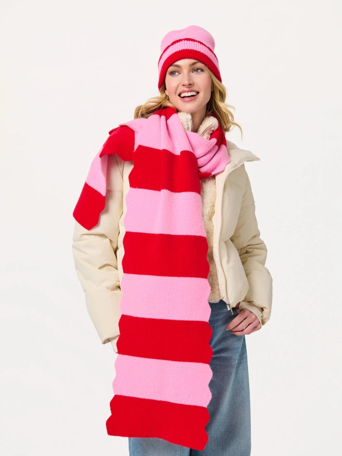 A woman wearing a cream puffer jacket, blue jeans, a pink and red striped beanie, and a long pink and red striped knit scarf, standing against a plain white studio background.