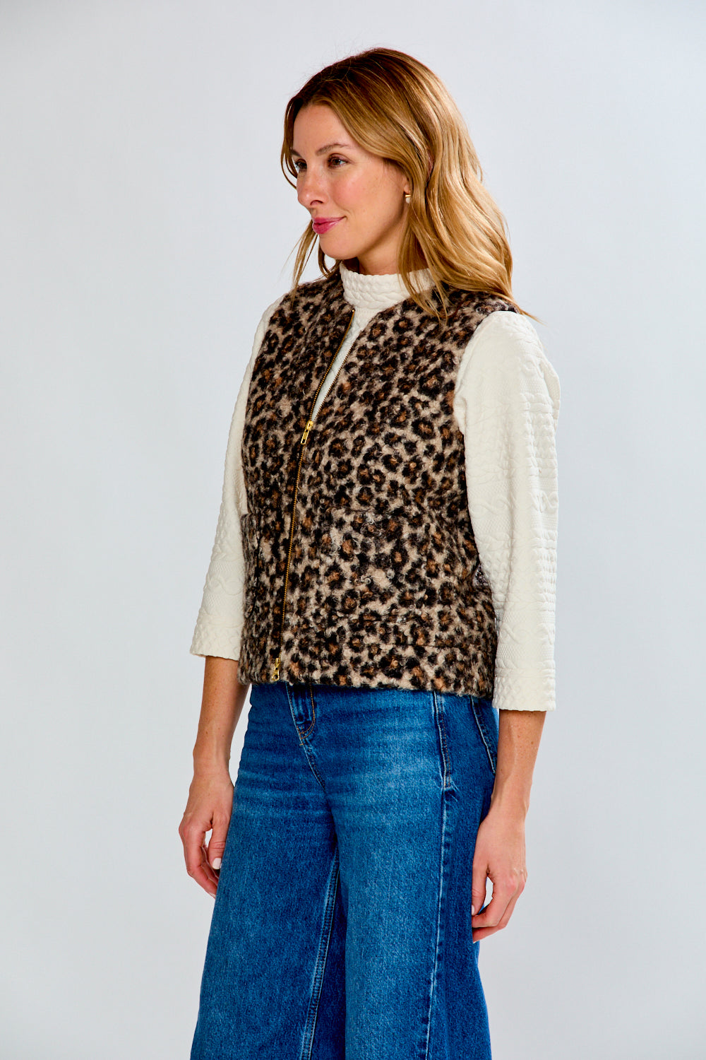Woman wearing a leopard print vest over a cream sweater with blue jeans on a white background