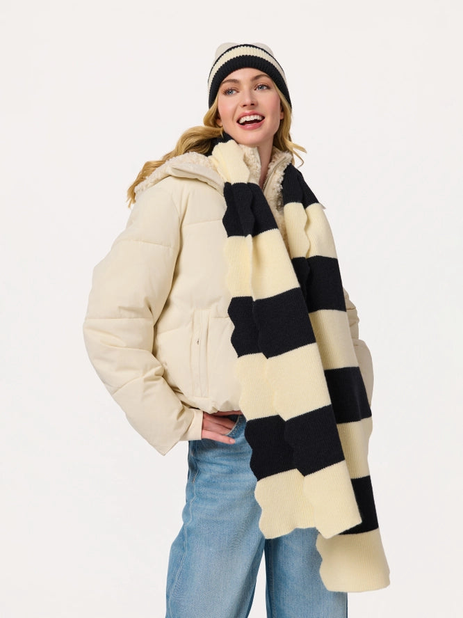 A woman modeling a cream puffer jacket, a cream and black striped knit scarf, and a cream and black striped beanie against a plain white studio background.