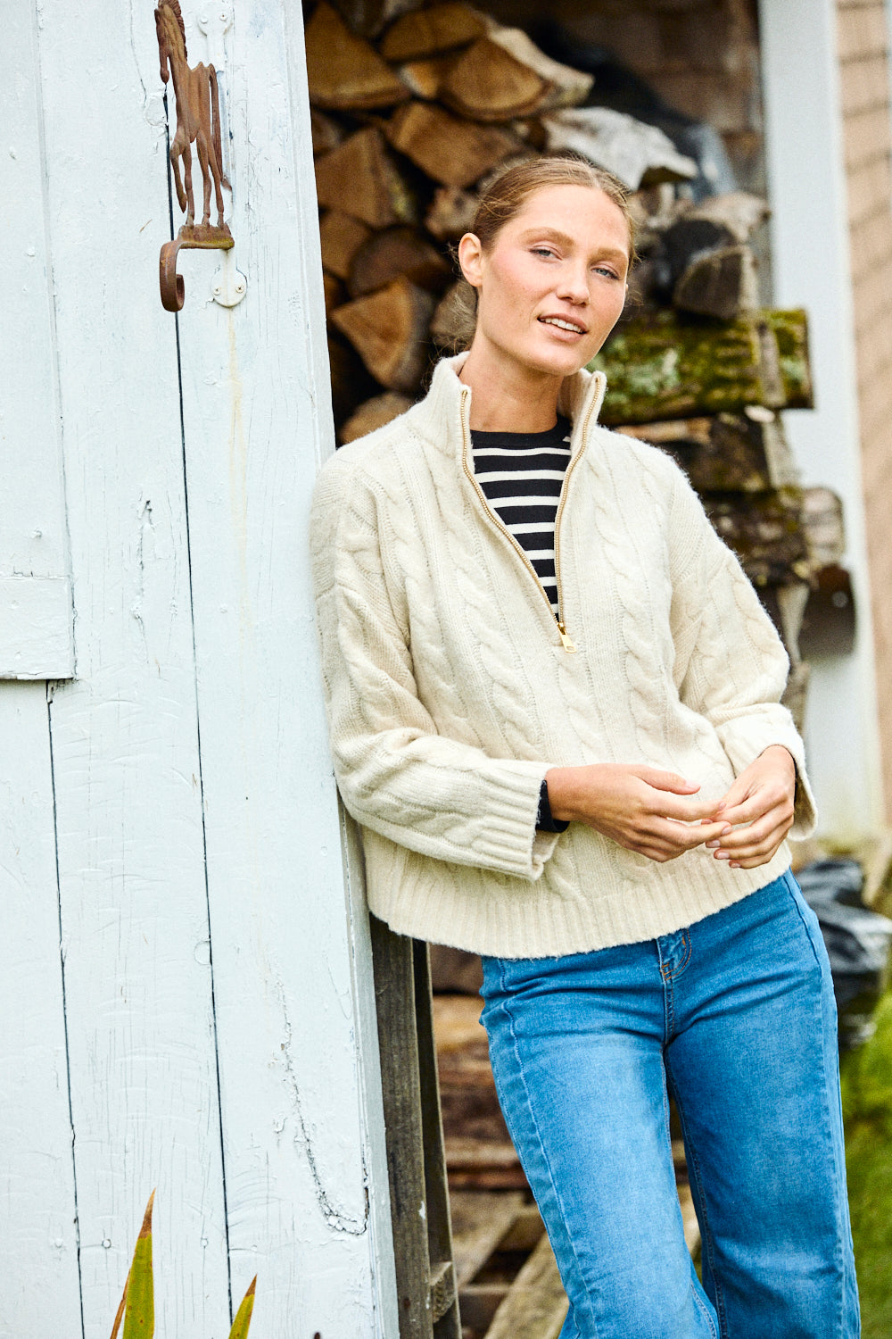 Woman wearing a cream jacket and blue jeans standing in front of a wooden shed.