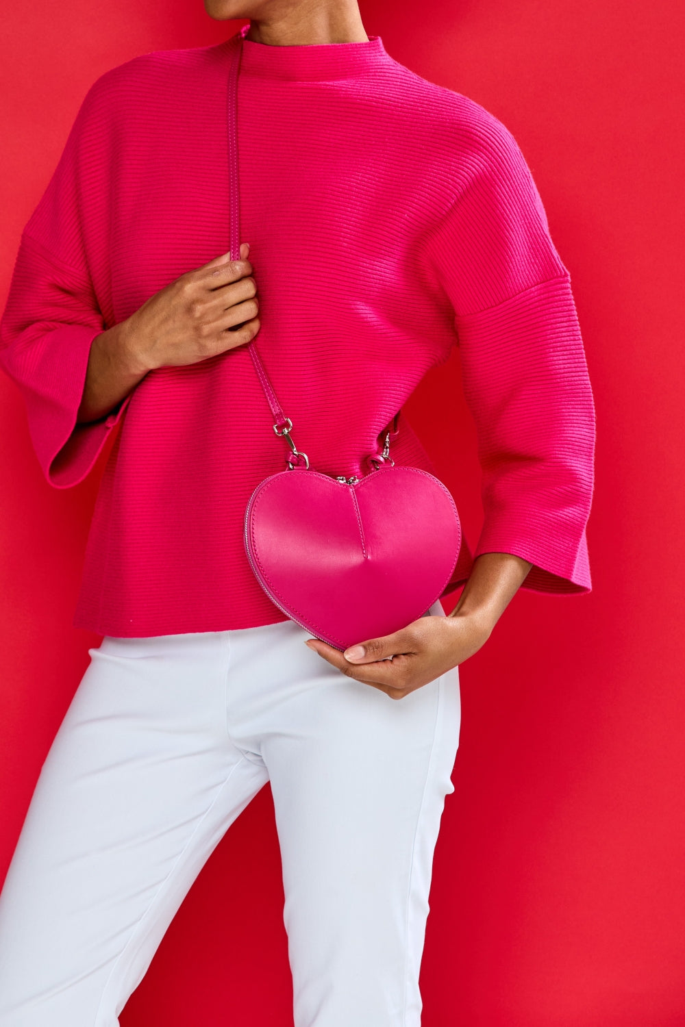 Person wearing a bright pink sweater and holding a matching heart-shaped handbag against a red background