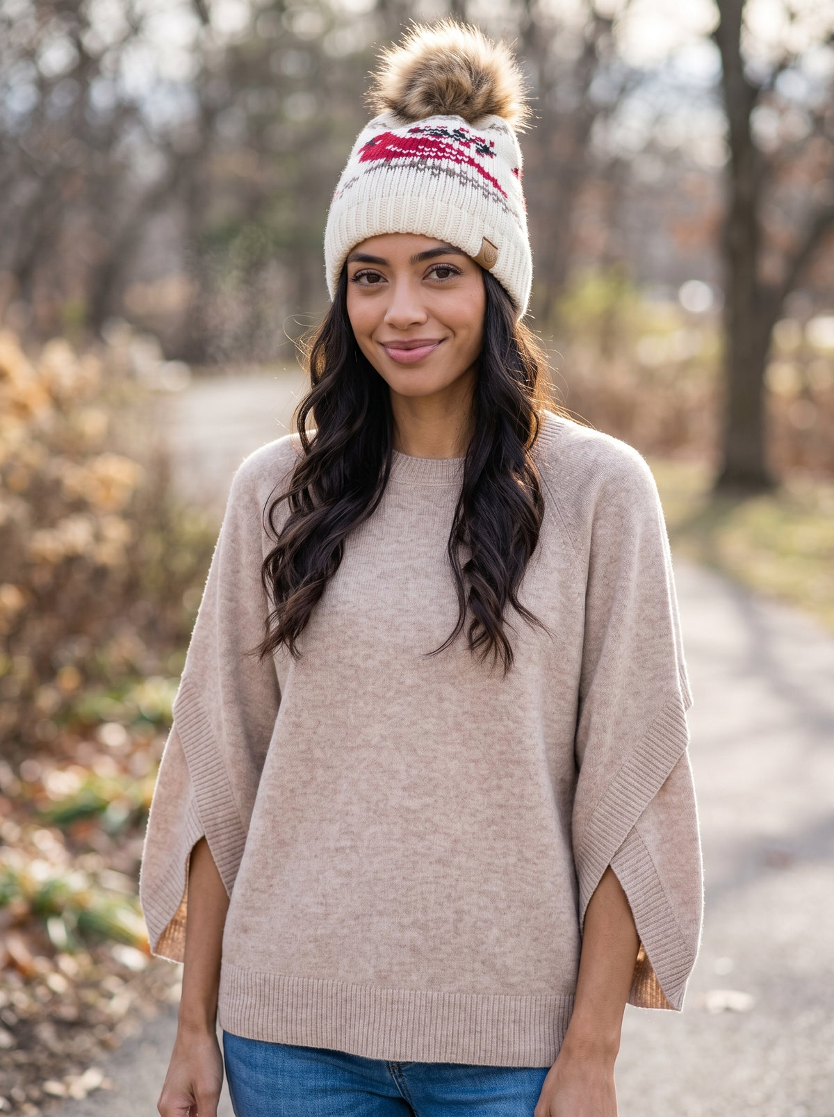 A woman wearing a beige knit poncho sweater and a cream knit beanie with a brown pom-pom and red pattern, standing on an outdoor path.
