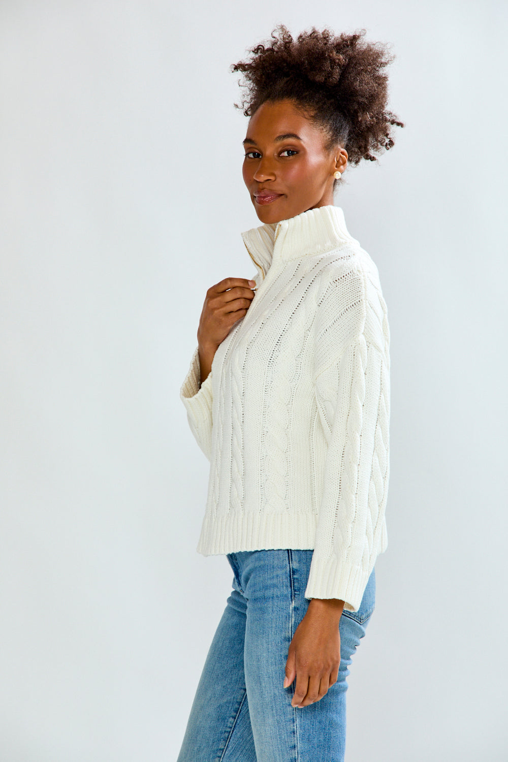 Woman wearing a white turtleneck sweater and blue jeans against a plain background