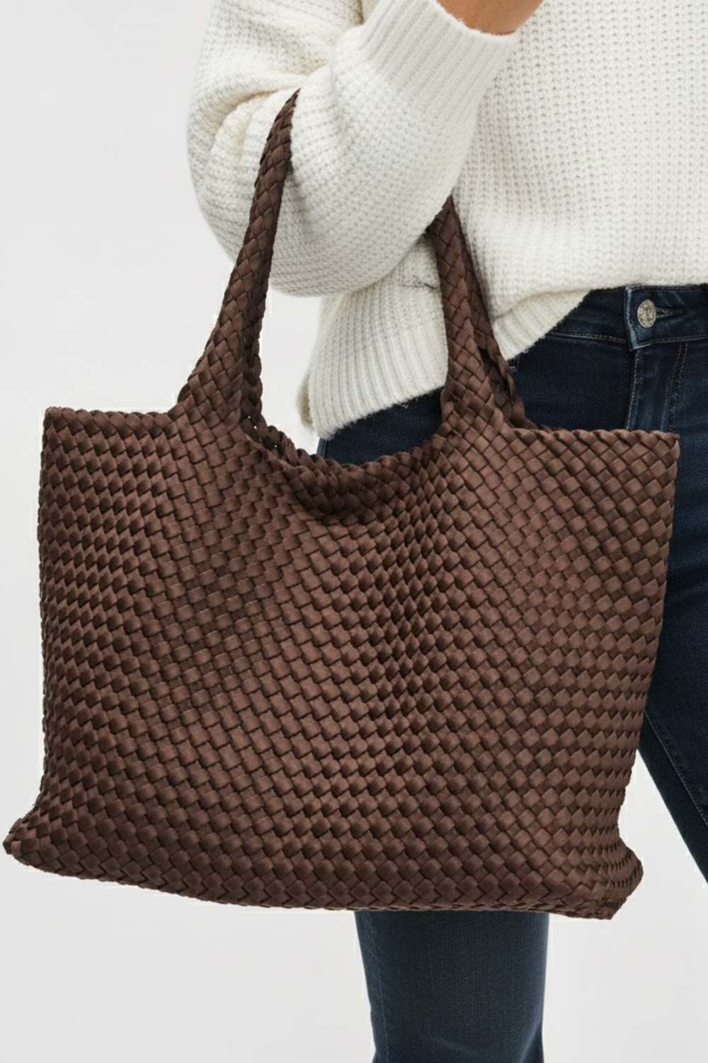 Brown woven tote bag held by a person wearing a white sweater and blue jeans on a light gray background