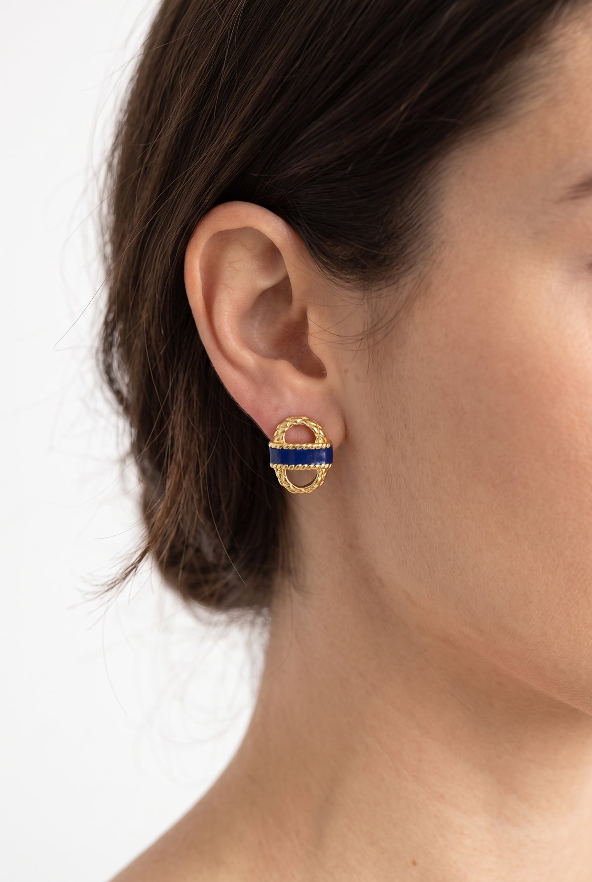 A close-up of a model's ear wearing a gold hoop earring with a central blue enamel band and small gold details.