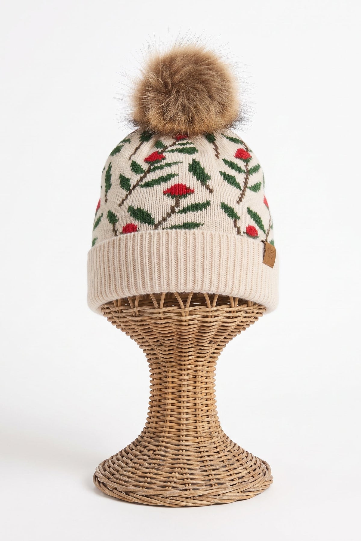 A knit beanie hat with a brown faux-fur pom-pom, featuring a cream base with a green and red floral pattern, displayed on a woven wicker stand against a white background.