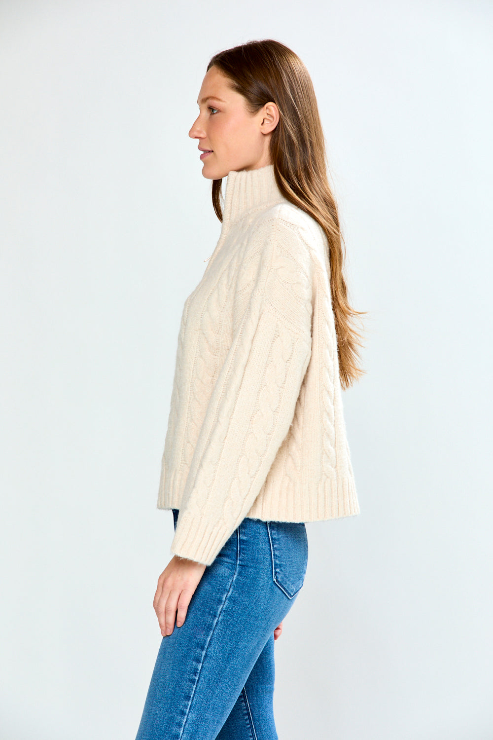 Woman wearing a oatmeal cable knit sweater and blue jeans on a white background