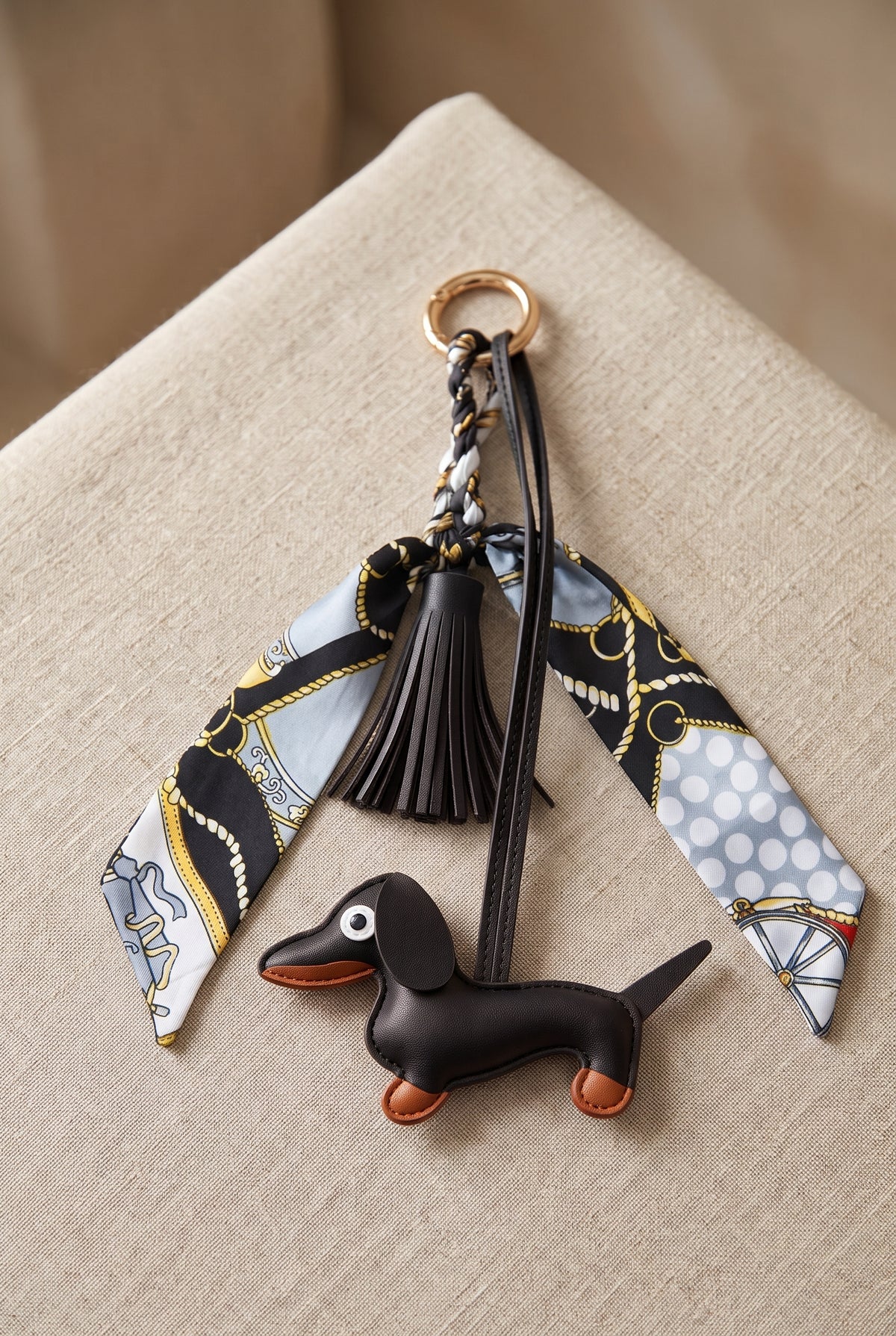 A fashion keychain featuring a black leather dachshund charm, a black leather tassel, a gold-tone key ring, and two patterned silk neckties in black, white, and gold, displayed on a beige textured fabric background.
