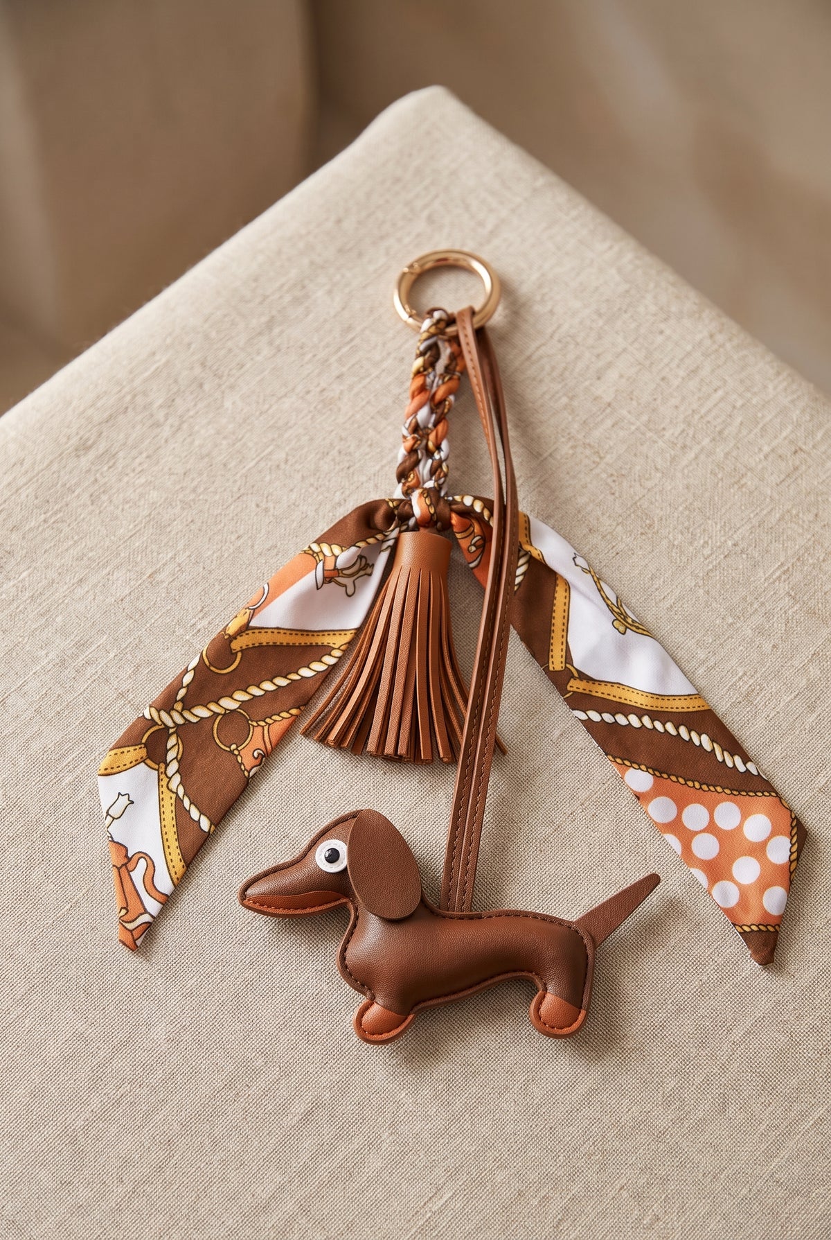 A brown leather dachshund-shaped keychain charm with a matching brown leather tassel and a patterned silk scarf wing, displayed on a beige textured fabric background.