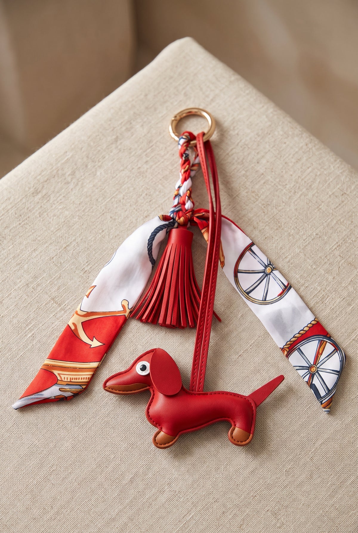 A red leather dachshund-shaped keychain charm with a red tassel, a gold key ring, and two patterned silk scarf ribbons, displayed on a beige textured surface.