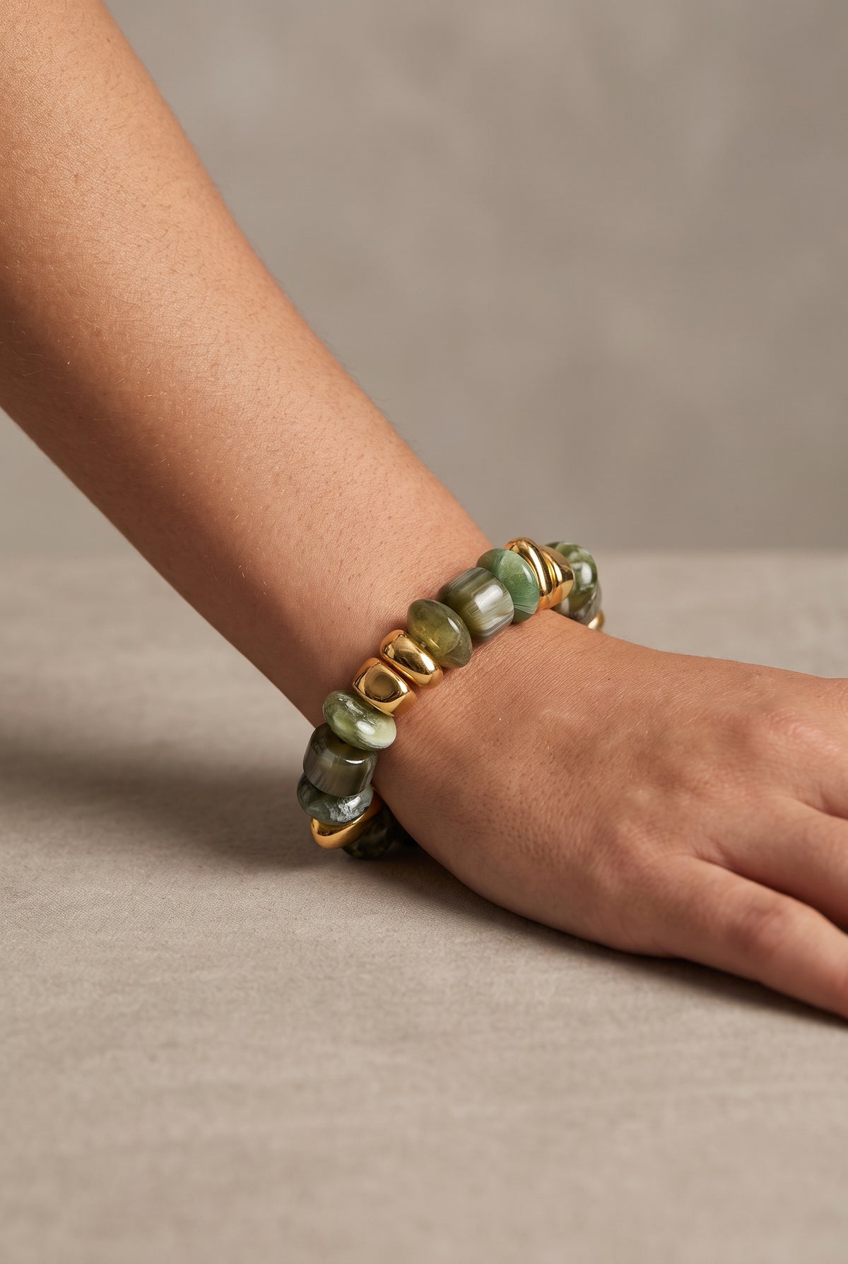 A close-up of a person's wrist and hand wearing a stretch bracelet made of polished green stone beads separated by small gold spacer rings, set against a neutral beige background.