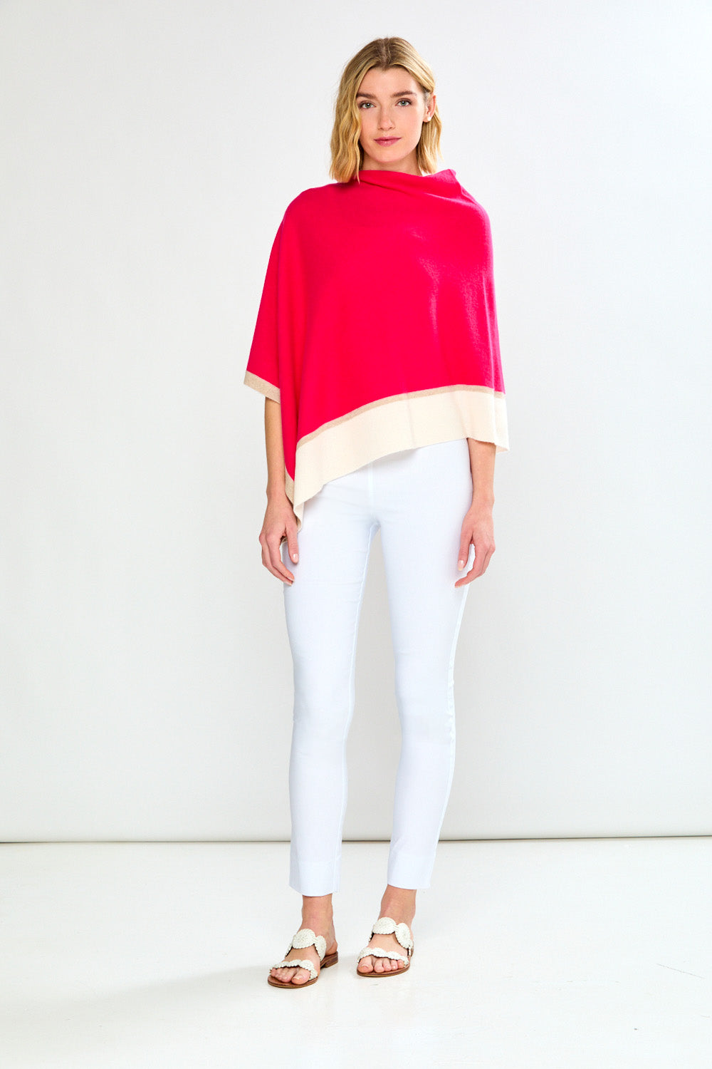 Woman wearing a bright pink poncho over a beige top with white pants on a white background