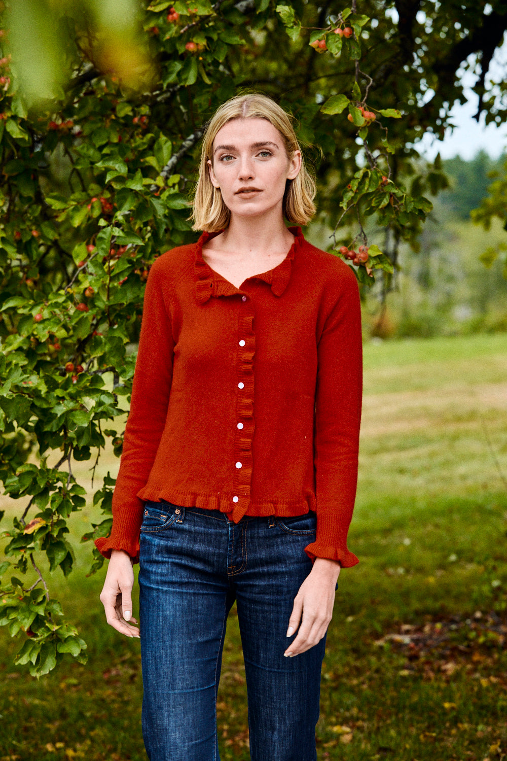 Woman wearing a red sweater and blue jeans standing in a natural setting with trees and grass.