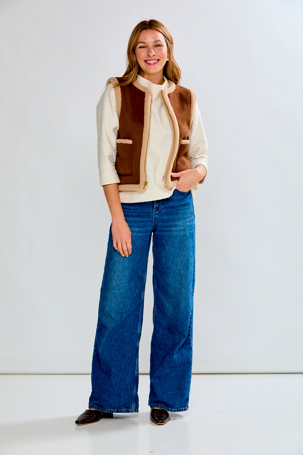 Woman wearing a brown and beige vest over a white shirt with blue jeans on a white background