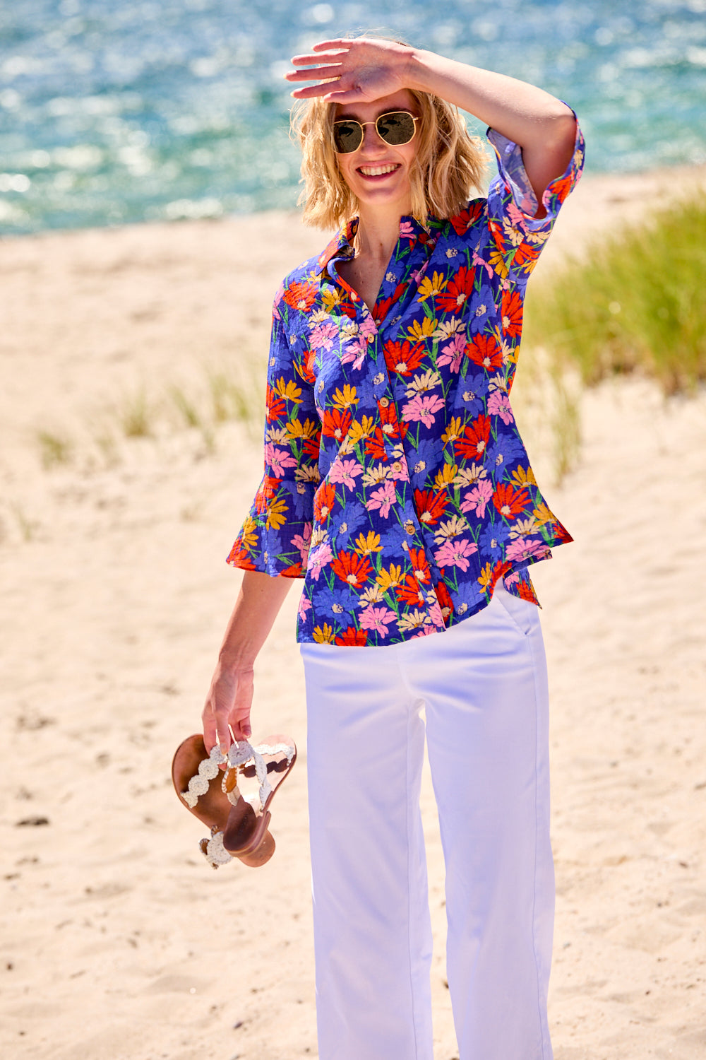 Woman in a colorful floral shirt and white pants standing on a beach.