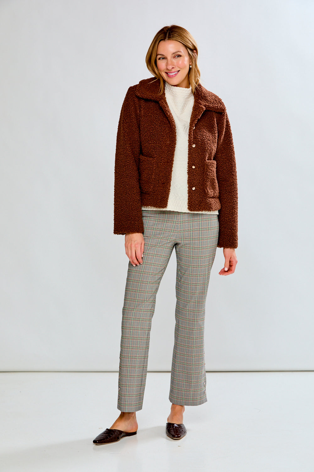 Woman wearing a brown jacket, white sweater, and plaid pants on a white background