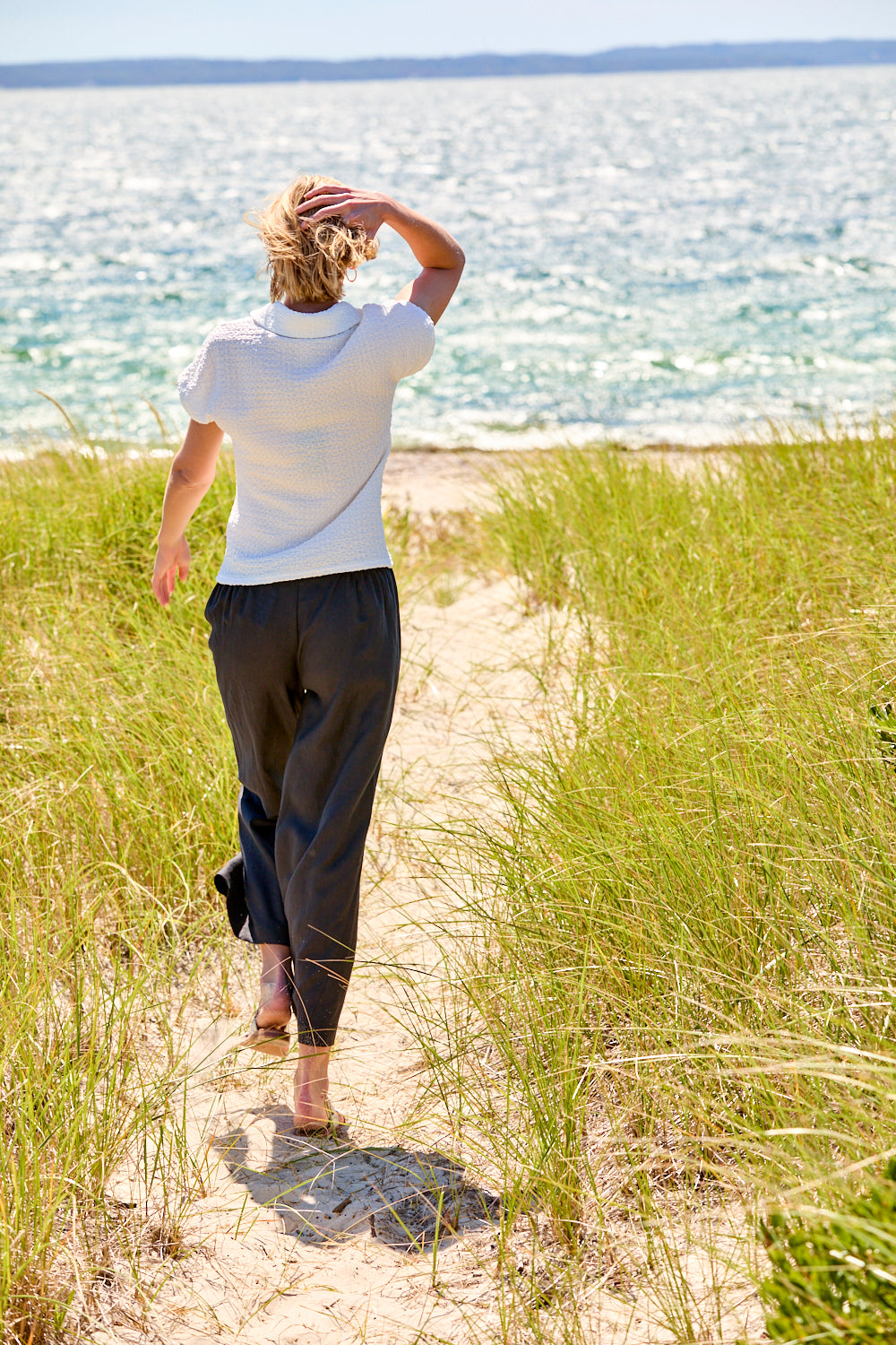 Person walking along a sandy path with grass on either side, looking out towards the ocean.