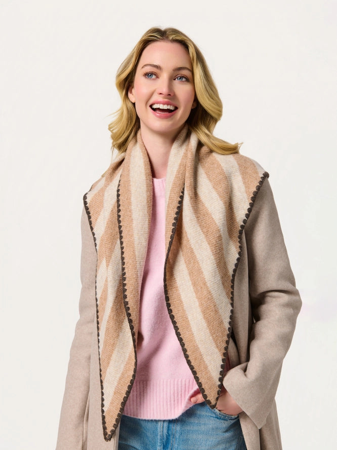 A woman wearing a beige and cream striped knit scarf with dark brown pom-pom trim, styled with a light beige cardigan and a pink sweater against a plain white background.